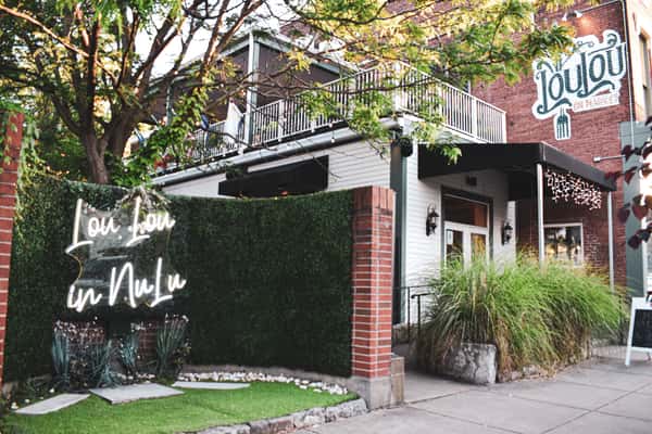 Front exterior of Lou Lou on Market, showcasing the signature "Lou Lou in NuLu" neon sign, lush greenery wall, and upstairs balcony—setting the tone for stylish, music-forward events in Louisville's NuLu district.