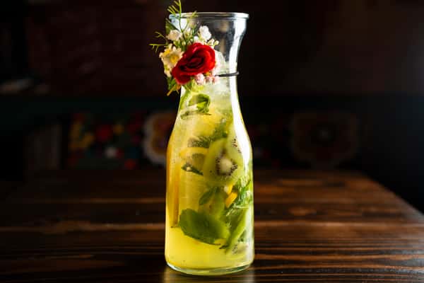A refreshing cocktail in a glass pitcher, featuring mint, kiwi, and lemon slices, garnished with a red rose and white flowers, set against a dark wooden background.