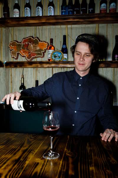 A bartender pours red wine into a glass, showcasing a cozy bar with various bottles in the background, highlighting the cultural ambiance of the establishment.