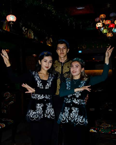 Three individuals dressed in traditional Central Asian attire pose together, showcasing intricate patterns on their outfits, with colorful mosaic lanterns illuminating the background.