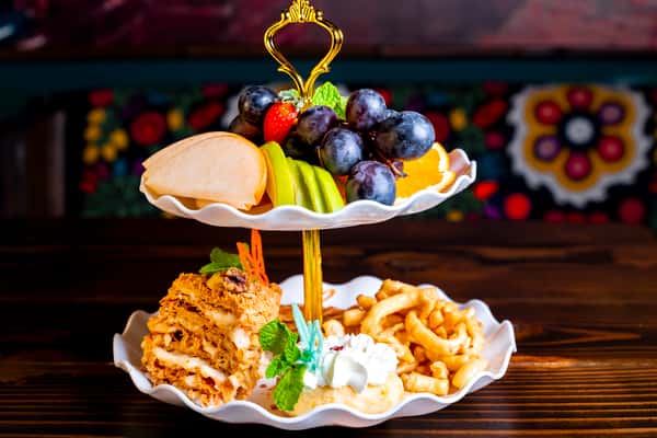 A two-tiered dessert platter featuring a variety of fresh fruits like grapes, apples, and oranges, alongside a layered cake and sweet pastries, garnished with mint.