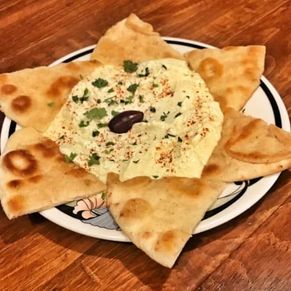 Creamy dip garnished with paprika, parsley, and a Kalamata olive, served with warm pita bread slices on a small plate.