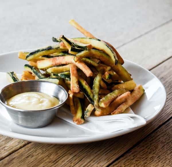 Crispy zucchini fries served on a white plate with a side of creamy dipping sauce in a small metal cup.