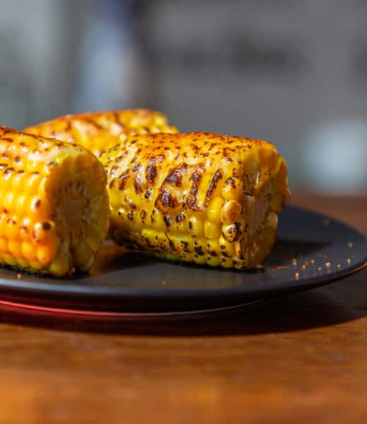 Grilled corn on the cob with char marks, served on a dark plate and lightly seasoned with chili powder.