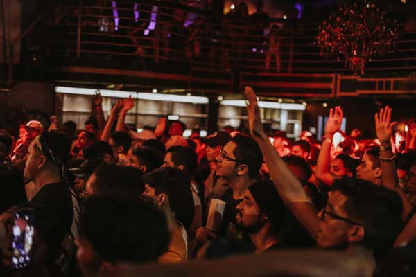 A large, diverse crowd of people dancing and raising their hands at a nightclub, lit with red and purple lights.