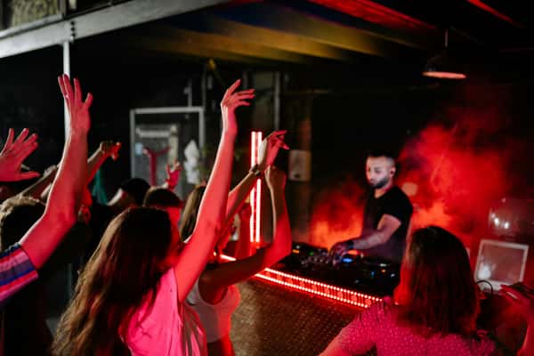 People dancing with hands raised at a nightclub, with a DJ and red lights in the background.