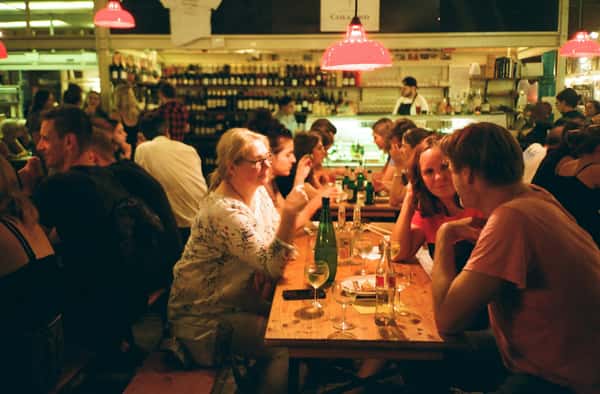 Diners seated at wooden tables in a dimly lit restaurant with a bar featuring bottles and a bartender in the background.