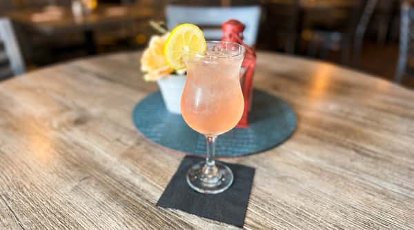 Light pink cocktail with ice and a lemon slice garnish, on a wooden table with a black napkin.