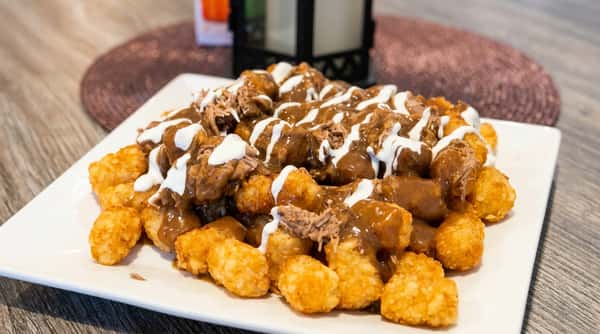 Tater tot poutine topped with shredded beef, rich brown gravy, and white cream sauce on a white plate.