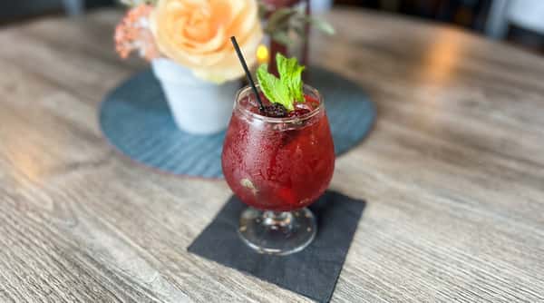 Red cocktail with blackberries, mint sprig, and black straw on a wooden table.