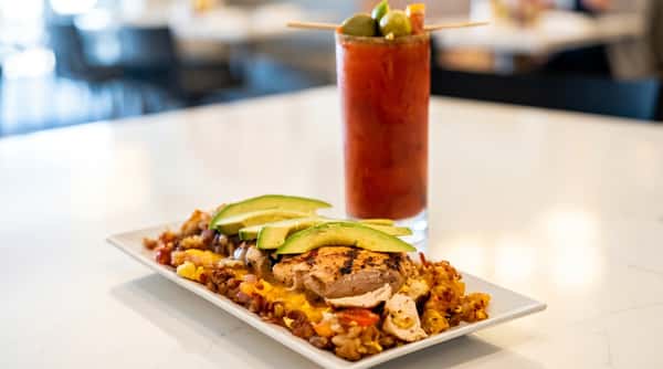 Grilled chicken and rice dish topped with melted cheese and sliced avocado, with a Bloody Mary in the background.
