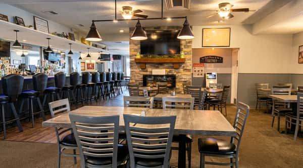 Restaurant interior with a stone fireplace under a TV, bar seating with dark stools, and tables with gray chairs.