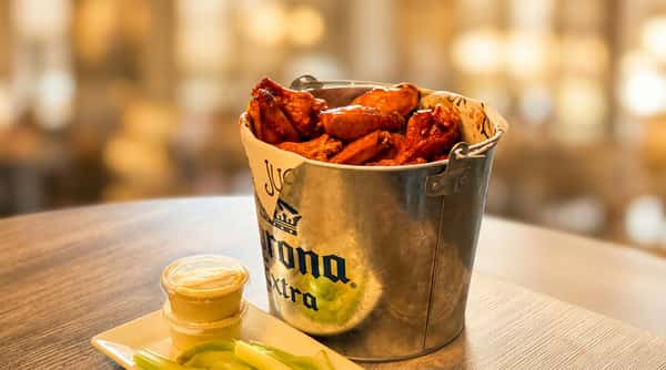Bucket of glazed chicken wings with celery sticks and dipping sauces on a striped wooden table.