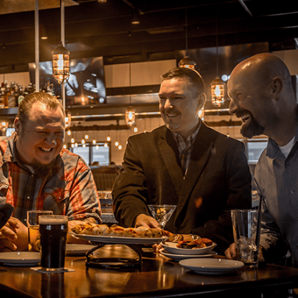 Group of smiling men enjoying drinks and shared appetizers at a lively bar, creating a fun and social dining atmosphere.