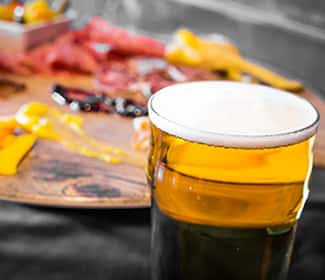 A frosty pint of golden beer with a creamy head, served alongside a charcuterie board featuring assorted meats, cheeses, and dipping sauces in the background.