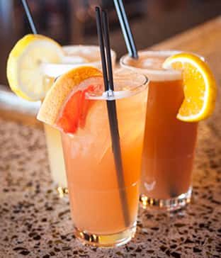 Refreshing fruit cocktails garnished with orange, grapefruit, and lemon slices, served in tall glasses with black straws on a granite bar counter.