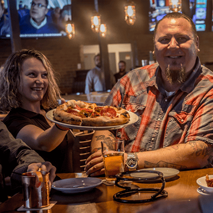 Smiling diners enjoying drinks at a lively bar, with a woman serving a freshly baked pizza topped with pepperoni and sausage.