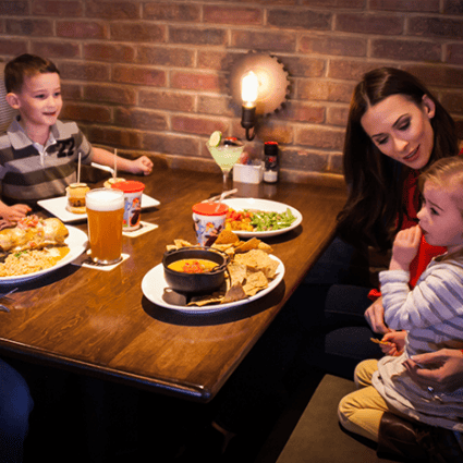 A family enjoying dinner at a cozy restaurant, with plates of nachos, salads, and kids' meals on a wooden table, set against a warm brick wall backdrop.