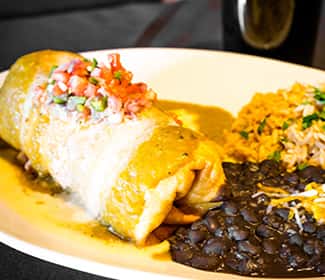 A hearty burrito topped with melted cheese and fresh pico de gallo, served with seasoned rice and black beans on a white plate.
