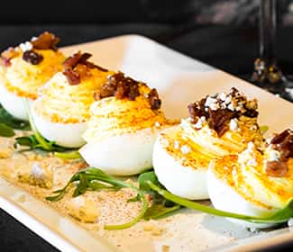 Creamy deviled eggs topped with crispy bacon bits and crumbled blue cheese, served on a bed of fresh greens on a rectangular white plate.