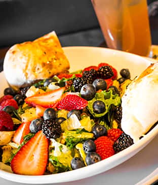 Fresh mixed greens salad topped with strawberries, blueberries, blackberries, and raspberries, served with two pieces of toasted bread and a drink in the background.