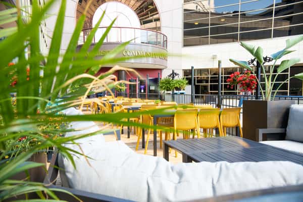 Outdoor patio seating at The Spoke café and bar with yellow chairs, lounge seating, and lush plants in a bright urban courtyard.
