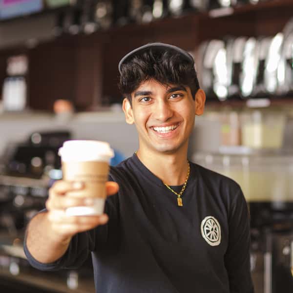 Smiling barista holding a takeaway coffee cup inside The Spoke café, kitchen, and bar.