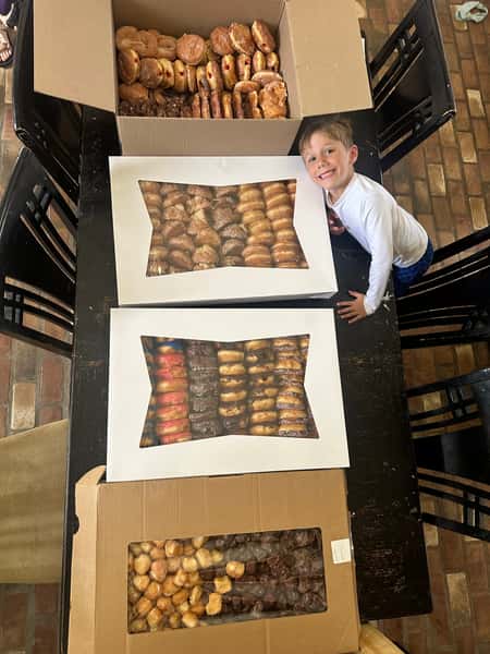Boy smiling next to large boxes of assorted donuts on a dining table.