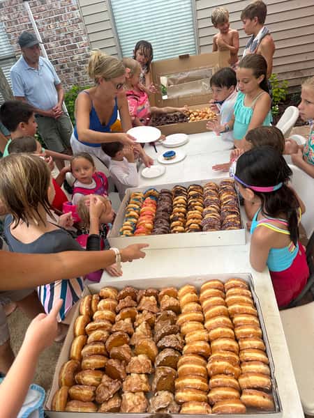Children gathered around tables filled with assorted donuts at an outdoor party, as a woman serves plates.