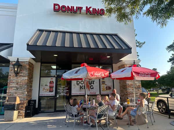 Family enjoying donuts and drinks at an outdoor table in front of Donut King, under Coca-Cola umbrellas on a sunny day.