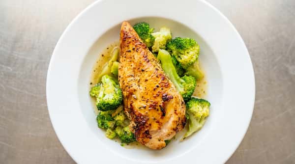 Grilled chicken breast with herbs and glazed sauce, served with steamed broccoli florets in a white bowl.