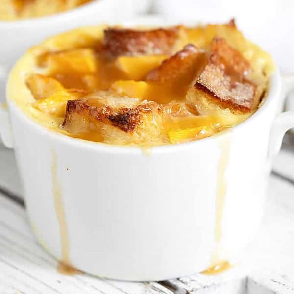 This is a serving of peach bread pudding in a white ramekin, topped with golden toasted bread cubes and drizzled with a rich caramel or butterscotch sauce.