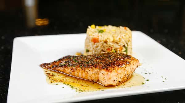 Pan-seared salmon fillet with seasoning, butter sauce, and a cylinder of vegetable fried rice on a white plate.