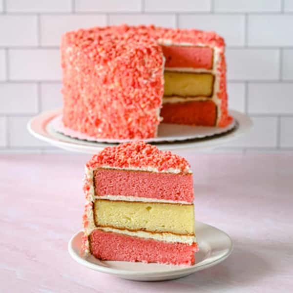 A three-layer strawberry crunch cake with alternating pink and yellow layers, filled and frosted with cream, and coated in a pink crumb topping. One slice is served on a small plate in front of the whole cake.