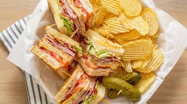 Club sandwiches with lettuce, tomato, bacon, and ham, served with a side of crinkle-cut potato chips and pickles.