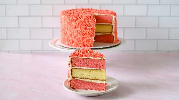 Strawberry crunch cake with alternating pink and yellow layers, covered in pink crumble and white frosting. A slice is in front.
