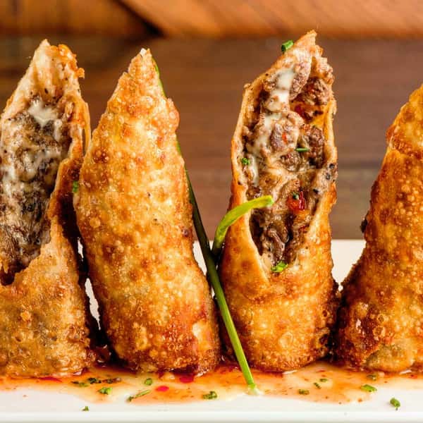 Crispy cheesesteak egg rolls filled with seasoned beef, melted cheese, and diced vegetables, served on a plate with a drizzle of sweet chili sauce.