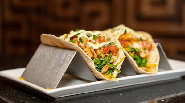 Two tacos with diced chicken, lettuce, tomato, and a creamy sauce in a metal holder.