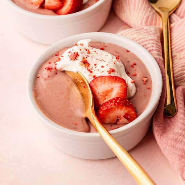 Strawberry mousse served in a white ramekin, topped with whipped cream and fresh strawberry slices, with a gold spoon.