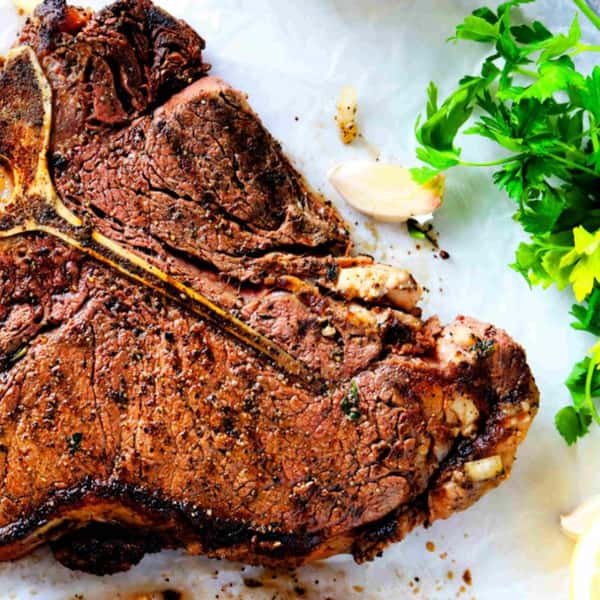 Grilled T-bone steak seasoned with black pepper and herbs, served with garlic cloves and fresh parsley on the side.