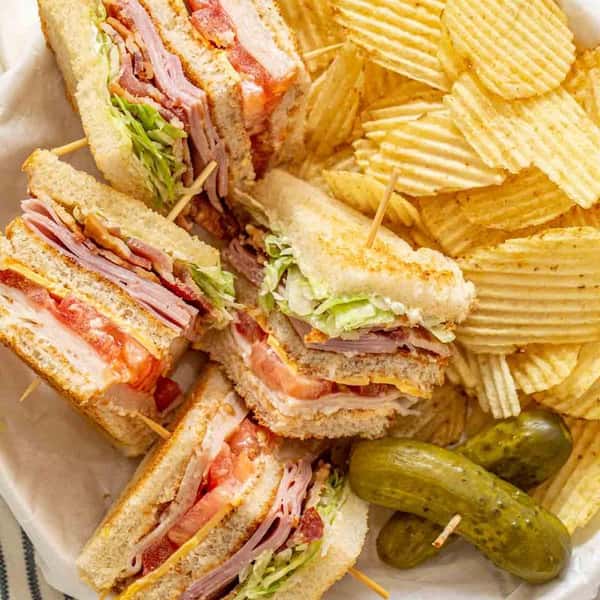 This image shows a classic club sandwich platter, featuring stacked club sandwiches filled with deli meats, cheese, lettuce, tomato, and bacon, served with ridged potato chips and a few whole pickles on the side.