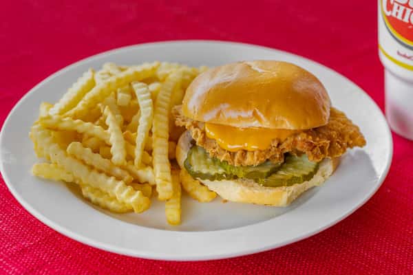 Crispy chicken sandwich with cheddar cheese and pickles on a glossy toasted bun, served alongside crinkle-cut fries on a white plate over a red textured tablecloth.