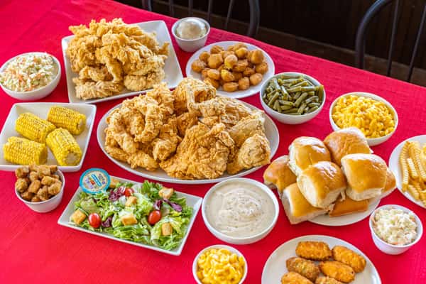 Hearty Southern comfort food feast featuring crispy fried chicken, chicken tenders, fluffy dinner rolls, hush puppies, crinkle-cut fries, mac and cheese, green beans, coleslaw, corn on the cob, fried okra, a fresh garden salad with buttermilk ranch, and creamy white gravy, all beautifully arranged on a vibrant red tablecloth.