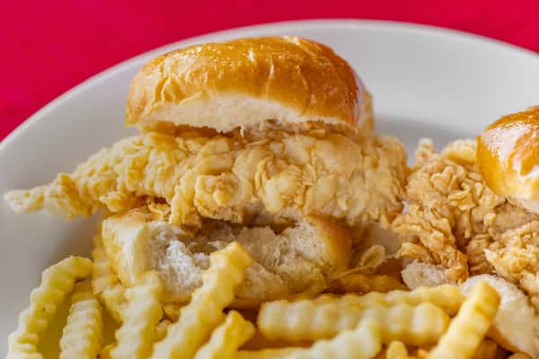 Close-up of a golden chicken tender roll from Bush's Chicken, featuring a crispy fried chicken tender nestled in a soft, shiny bun, served with crinkle-cut fries on a white plate.