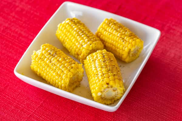 Buttered corn on the cob, seasoned with black pepper, served in a white square bowl on a red tablecloth—classic Southern side dish.