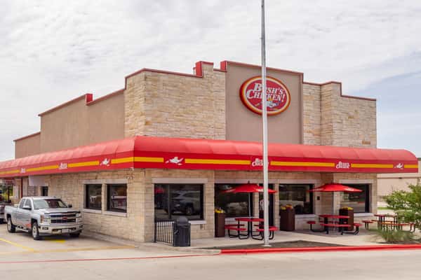 Exterior of a Bush's Chicken restaurant featuring tan brick walls, a bold red awning with yellow accents, outdoor seating with red umbrellas, and a pickup truck parked in front under a partly cloudy sky.