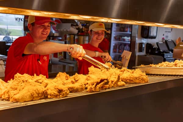 Bush's Chicken employees smiling while serving freshly fried chicken from behind the counter, showcasing a golden batch of crispy chicken under heat lamps in a welcoming fast-food kitchen setting.