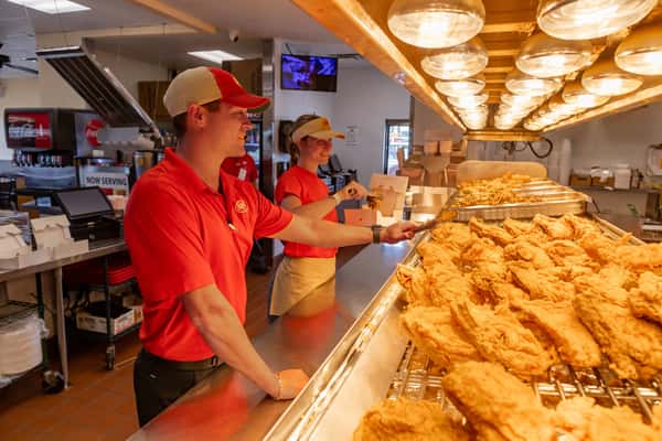 Bush's Chicken employees smiling behind the counter while serving fresh, golden fried chicken from a well-lit heat lamp display in a welcoming fast-food kitchen environment.