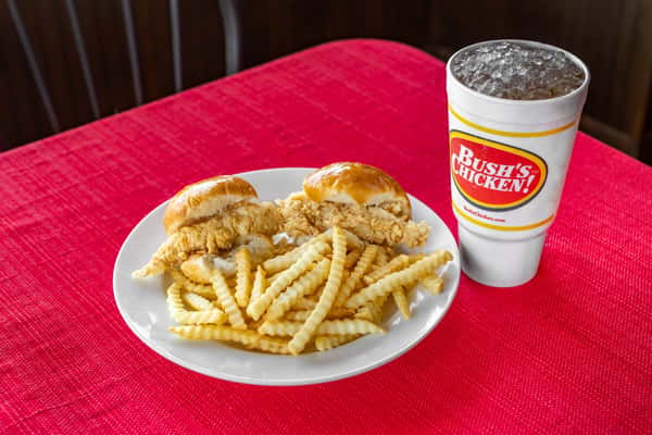 Chicken tender roll meal from Bush's Chicken featuring two soft rolls filled with crispy chicken tenders, served with crinkle-cut fries and a large iced drink on a white plate over a red tablecloth.