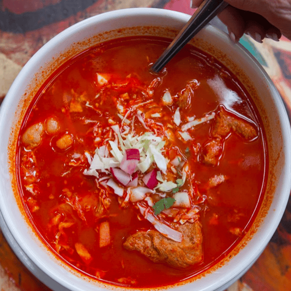 Bowl of red pozole topped with shredded cabbage, diced radish, onion, and tender chunks of pork.